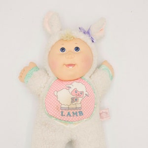 May include: A white Cabbage Patch Kid doll wearing a white lamb costume with pink and green accents. The costume has a pink bib with a white lamb illustration and the word "LAMB" printed on it.