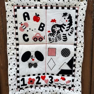 May include: A black and white baby play mat with a red, white, and black border. The mat has four squares with different designs. The top left square has the letters A, B, and C, a red apple, a bee, and a red car. The top right square has a zebra and a smaller zebra. The bottom left square has a black and white dog with a red bow tie. The bottom right square has a red square, a black circle with white dots, a white diamond, and a black triangle. The bottom row has the numbers 1, 2, and 3, a red heart, and two red fish.