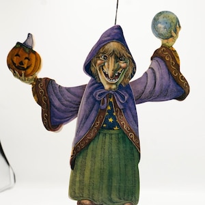 May include: A whimsical Halloween decoration featuring a smiling witch in a purple hooded cloak and green skirt. She holds a carved pumpkin and a globe, with gold accents on her sleeves and bodice. The witch is suspended by a black string.