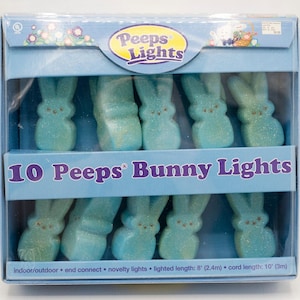May include: A box of 10 Peeps Bunny Lights. The lights are shaped like blue bunnies with glitter. The box has the Peeps Lights logo and text that reads "10 Peeps Bunny Lights". The box is blue and has a clear plastic front.