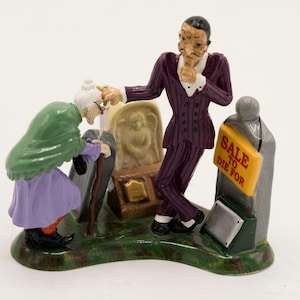 May include: A ceramic figurine depicting an elderly person with a cane and a tall figure in a purple pinstriped suit standing by a tombstone. The tombstone features a yellow sign with the red text SALE TO DIE FOR and a small angel relief.