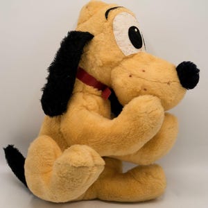 May include: A plush toy of Pluto, a cartoon dog from Disney. Pluto is sitting with his paws in front of him. He has a red collar and big, round eyes.
