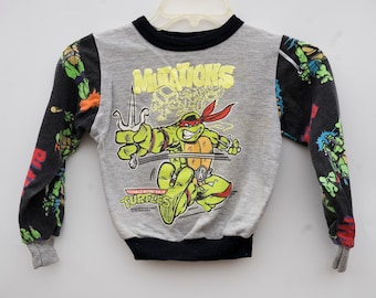 May include: A gray long-sleeve shirt with a black and green Teenage Mutant Ninja Turtles graphic. The shirt features a Teenage Mutant Ninja Turtle character holding a sai and the text "Mutations" and "Teenage Mutant Ninja Turtles".