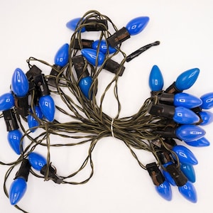 May include: A string of decorative holiday lights featuring multiple opaque blue, teardrop-shaped bulbs attached to a dark green electrical cord. The bulbs are arranged in a tangled cluster, each secured by a black plastic socket base.
