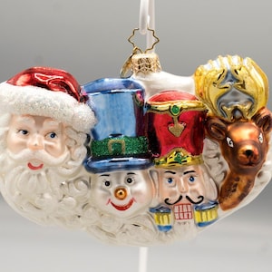 May include: A glass Christmas ornament featuring a horizontal arrangement of four figures: Santa Claus in a red hat, a snowman in a blue top hat, a nutcracker in a red uniform, and a brown reindeer with gold antlers, all set against a white base.