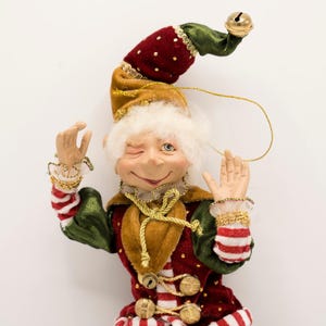 May include: A winking elf ornament with white hair, wearing a red and green velvet outfit with gold trim, metallic buttons, and striped sleeves. The elf has a long, pointed hat topped with a small gold bell and includes a thin gold hanging cord.