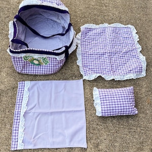 May include: A vintage doll carrier set in purple and white gingham check. The set includes a carrier, a blanket, a pillow, and a sheet, all trimmed with white lace. The carrier has the text "Cabbage Patch Kids" on the front.