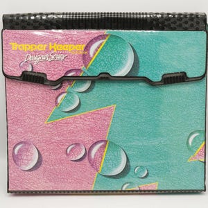 May include: A Trapper Keeper Designer Series notebook with a textured surface featuring a pink and teal geometric pattern with bubble graphics. The front flap has a black plastic closure and the text Trapper Keeper Designer Series printed in yellow.