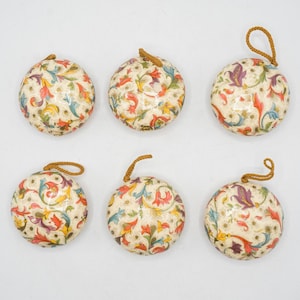 May include: Six round, flat ornaments featuring a cream-colored background with a repeating pattern of red, blue, yellow, and purple floral scrolls and small gold dots. Each ornament includes a small, braided brown cord loop for hanging.