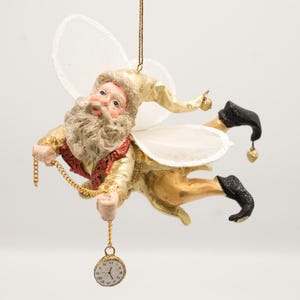 May include: A hanging ornament featuring a Santa Claus figure with white wings, wearing a gold hat and suit with a red vest. The figure holds a gold chain attached to a small round clock face with Roman numerals, and wears black pointed boots.