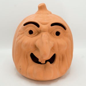 May include: A terracotta-colored pumpkin-shaped Halloween decoration with a carved face. The face features large, arched eyebrows, round eye holes, a long nose, and a wide, toothy grin. The pumpkin has a stem at the top and is designed to be a festive holiday accent.