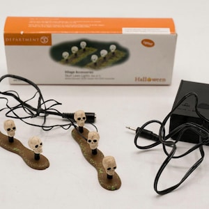 May include: A Department 56 Halloween village accessory set featuring two brown textured bases with three miniature off-white skulls each, connected by black wires to a black battery pack. The original orange and white box is included.