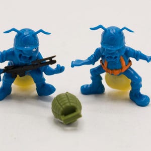 May include: Two blue toy figures with antennae and yellow bellies, one holding a black rifle, the other with an orange harness. A green grenade is in front of them. The toys are in a battle stance.