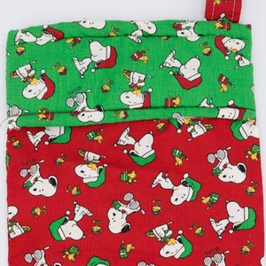 May include: Red and green Christmas stocking with a Peanuts theme. The stocking features a red background with white Snoopy characters and the words "Santa" and "Oops". The top of the stocking is green with white Snoopy characters.