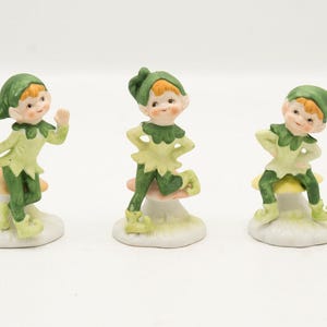 May include: Three ceramic elf figurines, each wearing a green hat and outfit with a pointed collar, sitting on individual yellow and white mushroom-shaped bases. The elves have light skin, orange hair, and are posed in different standing positions.