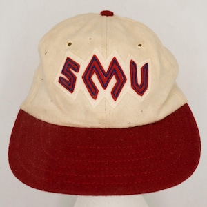 May include: Cream-colored baseball cap with a red brim and a red button on top. The cap features the letters "SMU" in a diamond-shaped design, with red letters outlined in blue.