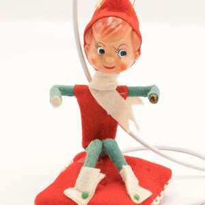 May include: A vintage Christmas elf ornament. The elf has a red felt outfit, a matching hat, and a white scarf. The elf is seated on a red pillow with a white lace trim. The elf's face is painted with rosy cheeks and blue eyes.
