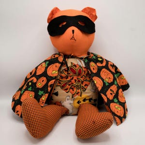 May include: Orange Halloween teddy bear wearing a black eye mask and a cape with a pumpkin pattern. The bear is sitting with its legs crossed and has a black and orange checkered pattern on its feet.