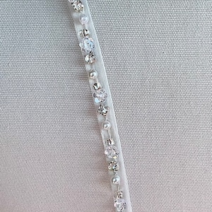 May include: A white ribbon with clear and pearl-like embellishments. The ribbon is beaded with small, clear crystals and white pearls.