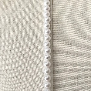 May include: A close-up of a white fabric with a row of white pearl beads sewn along the edge.