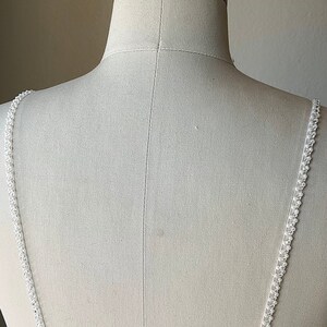 May include: White beaded straps for a wedding dress or other formal attire. The straps are made of small, white beads that are sewn together to create a delicate and elegant look.