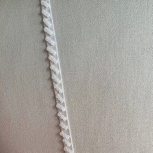May include: White beaded trim with a satin ribbon backing. The trim is shown on a light gray fabric background.