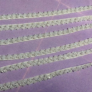 May include: White beaded trim with a sheer backing. The trim is arranged in four rows on a purple surface.