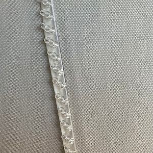May include: Close-up of a white fabric with a clear plastic trim. The trim has a beaded design.