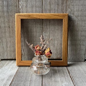 May include: A small, clear glass vase with dried flowers sits in front of a square, light brown wooden frame. The vase contains dried roses and lavender. The background is a weathered, gray wooden surface.