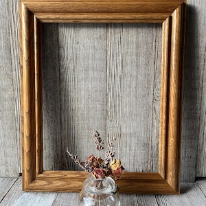 May include: A rectangular wooden picture frame with a light brown finish. The frame has a decorative edge and is empty. A small glass vase with dried flowers sits in front of the frame.