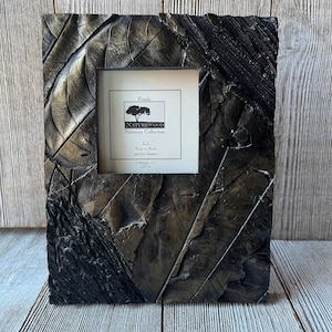 May include: A rectangular picture frame with a leaf design. The frame is black and bronze with the text "NATUREWOOD Polyresin Collection" and "5x5" on the included picture. The frame is made of polyresin.