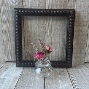 May include: A square black picture frame with a beaded edge, next to a small glass vase holding dried pink roses and lavender. The frame has a distressed finish, and the background is a wooden surface.