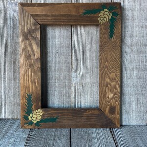 May include: A rectangular wooden picture frame with a medium brown stained finish. The frame features hand-painted details in the top right and bottom left corners, consisting of dark green pine needles and a gold-colored pinecone design.
