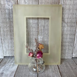 May include: A rectangular, translucent, pale yellow picture frame with a smooth finish, positioned behind a small, clear glass jar containing a bouquet of dried flowers, including pink rosebuds and sprigs of lavender with brown stems.