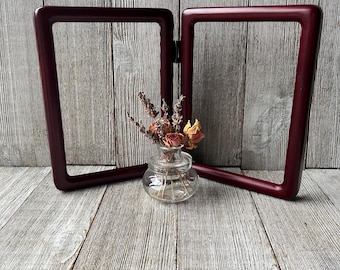 May include: A burgundy-colored, hinged double picture frame. The frame is empty, with a clear glass vase containing dried flowers in front of it. The background is a weathered, gray wooden surface.