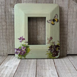 May include: A rectangular, light green picture frame with rounded corners. It features a butterfly and floral decals. The frame is decorated with purple violets and a butterfly with yellow and black wings. The frame is made of wood.