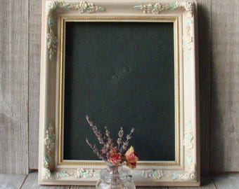 May include: A rectangular, cream-colored picture frame with ornate floral detailing and a gold inner border. The frame holds a dark green background. A small glass vase with dried flowers sits in front of the frame, adding a touch of vintage charm.