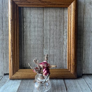 May include: A rectangular wooden picture frame with a medium brown finish and visible grain, positioned behind a small, clear glass jar containing a dried arrangement of pink rosebuds and sprigs of lavender with a thin metal wire handle.