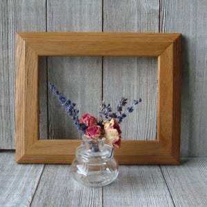 May include: A wooden picture frame with a simple design, featuring a small glass vase with dried flowers inside. The flowers are a mix of lavender and pink roses.
