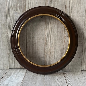 May include: A round, dark brown picture frame with a gold inner rim. The frame has a thick, rounded profile and a small hook on the back for hanging. The frame is empty, ready to display a photo or artwork.