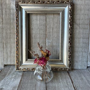 May include: A rectangular picture frame with a metallic silver finish and ornate, dark-toned scrollwork along the outer edge. In front of the frame sits a small, clear glass jar containing a dried arrangement of pink roses and lavender sprigs.
