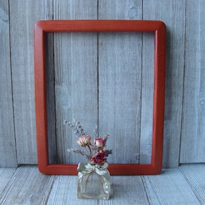May include: A wooden picture frame with a rounded edge, in a reddish-brown color, sits on a gray wood surface. A small glass vase with dried flowers is in front of the frame.