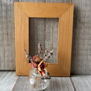 May include: A light brown wooden picture frame with a rectangular opening. A small glass jar with dried flowers and lavender sprigs sits in front of the frame. The background is a weathered wooden surface.