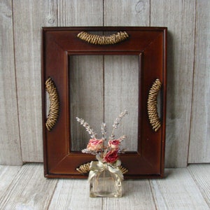 May include: A dark brown wooden picture frame with a woven design around the edges. The frame is empty and has a small vase with dried flowers in front of it.