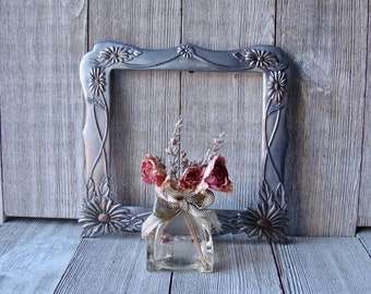 May include: A decorative silver-colored picture frame with floral accents, holding a small glass bottle filled with dried flowers and tied with a ribbon. The background is a weathered wooden surface.