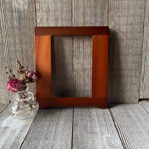 May include: A rectangular wooden picture frame with a smooth, reddish-brown finish and a wide border. Beside the frame sits a small, clear glass vase containing a cluster of dried pink roses and sprigs of lavender with thin, light brown stems.