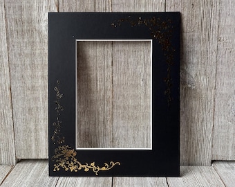 May include: A black picture frame with a white mat and gold floral corner and edge accents. The frame is rectangular with a central opening. The gold floral design adds a decorative touch to the frame.