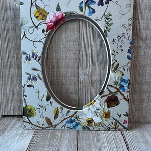 May include: A rectangular picture frame with an oval center opening. The frame features a light gray background decorated with a colorful botanical print of various flowers, including pink roses, blue blossoms, and white daffodils with green stems.