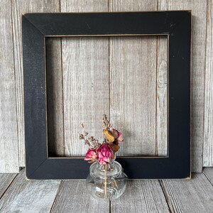 May include: A square black picture frame with distressed edges positioned behind a small clear glass jar containing a bouquet of dried pink roses and sprigs of lavender. The glass jar features a wire handle and embossed lettering on the base.