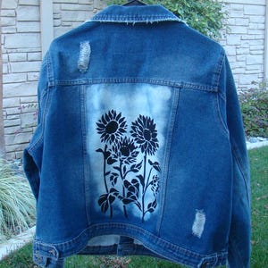 May include: A blue denim jacket with a distressed look and a white bleached area on the back. The bleached area features a black stencil design of sunflowers.